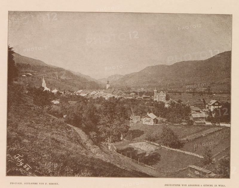View of Schladming, Östliche Ansicht von Schlädming, page, photomechanical print, number: 'IV.', photographer: Friedrich Simony, Salzburg (city), cliché maker: Angerer & Göschl, C., Vienna, publisher: Eduard Hölzel, 1888, Lower Austria, paper, autotypie, height 145 mm x width 195 mm, mountain-range, snow peaks, Steiermark, Schladming, A sepia-toned landscape photograph showing a valley with rolling hills and a village nestled between them, small clustered buildings and houses with pitched roofs spreading across the middle ground, a tall church tower and another steepled structure rising above the rooftops, open fields and fenced plots on the nearer slopes, a winding dirt path leading down the hillside, scattered trees and patches of vegetation across the scene, distant layered hills forming a backdrop under a broad, pale sky.