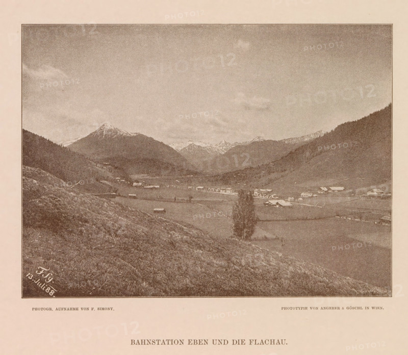 View of Flachau, Bahnstation eben und die Flachau, page, photomechanical print, number: 'XVII.', photographer: Friedrich Simony, Salzburg (city), cliché maker: Angerer & Göschl, C., Vienna, publisher: Eduard Hölzel, 1888-07-13, Lower Austria, paper, autotypie, height 142 mm x width 191 mm, city-view in general, 'veduta', Flachau, A sepia-toned landscape photograph framed by a thin dark border shows a wide valley flanked by sloping hills and distant snow-capped mountains under a mostly clear sky with a few clouds. In the valley there are scattered small buildings and farm structures with open fields and patches of trees; a single taller tree stands near the foreground. The foreground consists of a grassy, rocky slope leading down into the valley, and the image is mounted on a pale background with printed captions visible below the framed photograph.