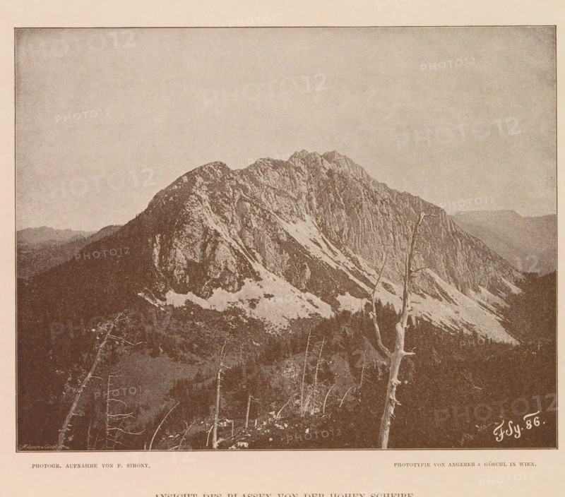 View of a mountain peak in the Dachstein Mountains, Ansicht des Blassen von der Hohen Scheibe, page, photomechanical print, number: 'XX.', photographer: Friedrich Simony, Dachstein (Salzburg), cliché maker: Angerer & Göschl, C., Vienna, publisher: Eduard Hölzel, 1886, Lower Austria, paper, autotypie, height 159 mm x width 200 mm, mountain-top, peak, A sepia-toned image showing a jagged mountain ridge with rocky faces and patches of snow, a foreground with bare tree trunks and branches, a darker wooded slope below the mountain, and a wide pale sky above, all framed by a thin border.