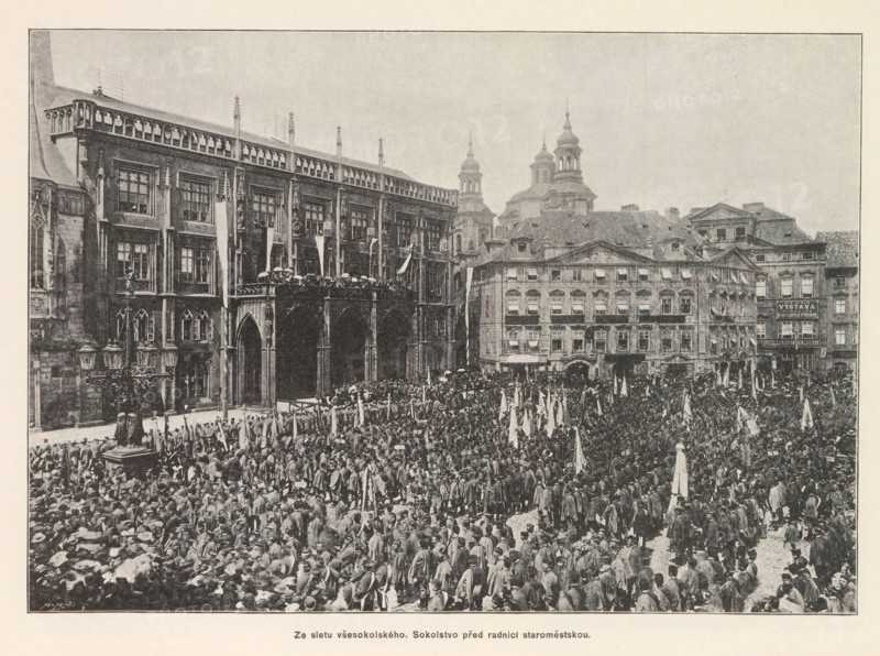 Crowd at the Staromestské námestí in Prague during a festival of the Sokol movement, Ze sletu vsesokolského. Sokolstvo pred radnici staromestskou, page, photomechanical print, Staromestské námestí, cliché maker: Carl Bellmann, Prague, c. 1891 - in or before 1893, photographer, paper, autotypie, height 187 mm x width 262 mm, public festivities, village square, Place, A large crowd fills a historic city square in front of ornate multi-story stone buildings with arched openings and decorative façades, many people holding upright banners and clustered closely together, a raised balcony area on one building crowded with people, several spired towers and domed roof structures visible behind the row of buildings, and a street lamp with ornate metalwork visible near the left side of the image.