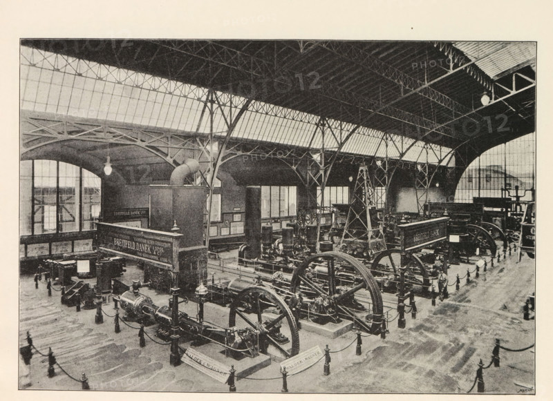 Instalace strojovny. Pohled smear k vychodnimu vchodu, Interior of an engine room, page, photomechanical print, Prague, cliché maker: Carl Bellmann, c. 1890 - in or before 1895, photographer, paper, autotypie, height 176 mm x width 258 mm, machines, parts of machines, tools and appliances, A large indoor industrial space with a high arched roof supported by metal trusses and columns, rows of tall windows along the upper walls, and hanging light fixtures. The floor area contains multiple large mechanical machines with visible flywheels, pistons, and belts, arranged on raised platforms and surrounded by short posts connected with chains. There are several tall structural towers or frames among the machinery and a few small display boards or panels mounted on walls. The overall scene is monochrome with a grainy texture and shows architectural details of the roof, beams, windows, and the dense arrangement of industrial equipment.