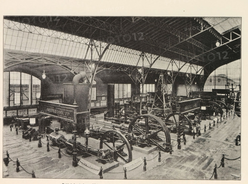 Pohled do strojovny. Výstava akciové strojirny drive Breitfeld, Danek a spol. v Katrine, Interior of a machine room with an exhibition of machines by Breitfeld and Danek, page, photomechanical print, Prague, cliché maker: Carl Bellmann, c. 1890 - in or before 1895, photographer, paper, autotypie, height 176 mm x width 258 mm, interior, representation of a building, machines, parts of machines, tools and appliances, A large indoor industrial hall with a high vaulted roof supported by metal trusses and arched iron columns, long rows of windows along the upper walls, hanging pendant lights, and a polished floor. Inside the hall are multiple large mechanical machines with prominent flywheels, pistons, and belts arranged on raised bases and connected by chains to low posts, with several decorative posts and ropes forming barriers around the exhibits. There are tall framed panels and a raised platform structure near one side, an overhead duct or pipe rising above a rectangular enclosure, and...