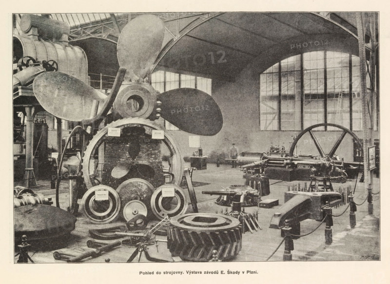 Pohled do strojovny. Výstava závodu F Ringhoffra na Smichove, Interior of an engine room with an exhibition of Skoda, page, photomechanical print, Prague, cliché maker: Carl Bellmann, c. 1890 - in or before 1895, photographer, paper, autotypie, height 176 mm x width 258 mm, interior, representation of a building, machines, parts of machines, tools and appliances, A large industrial interior with tall windows and arched roof supports contains heavy machinery and large metal components arranged across the floor; a prominent multi-bladed propeller is mounted near the center surrounded by circular gears and ring-shaped parts, with various shafts, pistons, and mechanical housings visible nearby; columns and structural beams rise toward the ceiling, and a large flywheel sits toward one side; workbenches, stands, and scattered smaller tools occupy the foreground and background, and the scene is rendered in a monochrome, grainy photographic style.