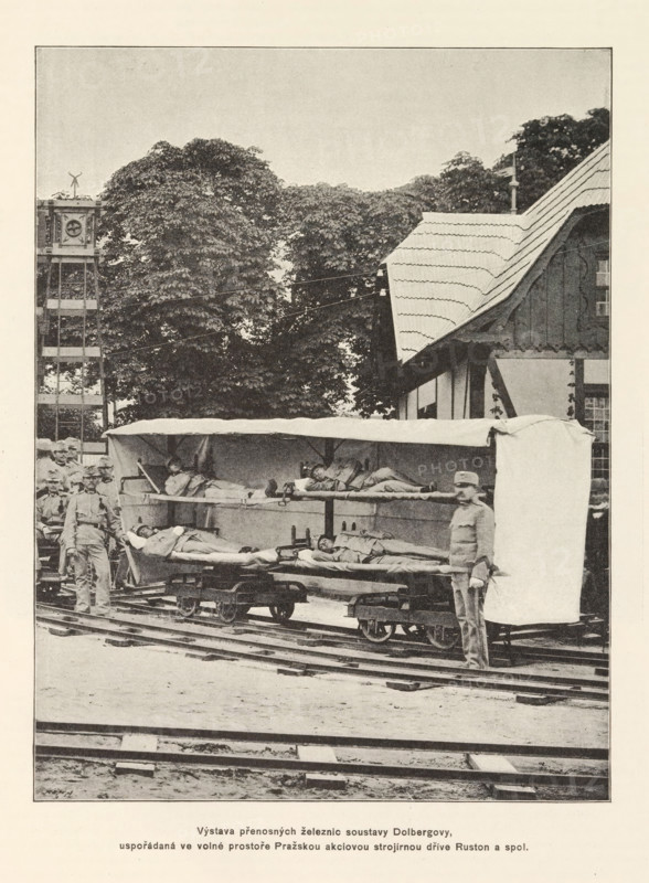 Sleeping compartment on rails, Výstava prenosných zeleznic soustavy Dolbergovy, uspodaradaná ve volné prostore Prazskou akciovou strojirnou drive Ruston a spol, page, photomechanical print, Czech Republic, cliché maker: Carl Bellmann, Prague, c. 1891 - in or before 1895, photographer, paper, autotypie, height 222 mm x width 171 mm, bed, railway-coach, railway-carriage, wagon, A sepia-toned photograph showing a rail setting with tracks in the foreground and a covered wagon-like stretcher platform on small rail carts holding people lying down under a canopy; several standing men in uniform are positioned around the platform, one near the front partially obscuring the end of the canopy, and others grouped toward the left side; behind them a timber-framed building with a steep, shingled roof occupies the right background while large leafy trees fill the central background; to the left a tall wooden tower structure rises above the trees.