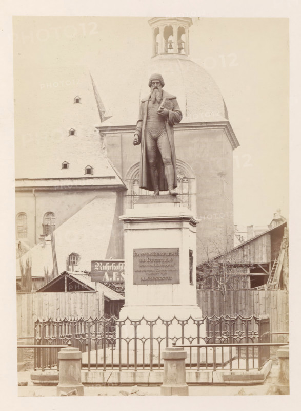 Monument to Johannes Gutenberg in Mainz, photograph, page, number: '15', photographer: B. Erdmann, Mainz, c. 1867 - in or before 1872, Rhineland-Palatinate, paper, albumen print, height 208 mm x width 150 mm, monument, statue, A tall stone pedestal stands behind a decorative metal fence, supporting a life-size bronze statue of a bearded man wearing a long coat and hat, with one hand holding a rolled object against his chest and the other hanging by his side, set against the façade of a large church with a steep roof, arched windows, and a domed bell tower visible above the statue, while wooden buildings and construction materials occupy the area around the base of the pedestal.