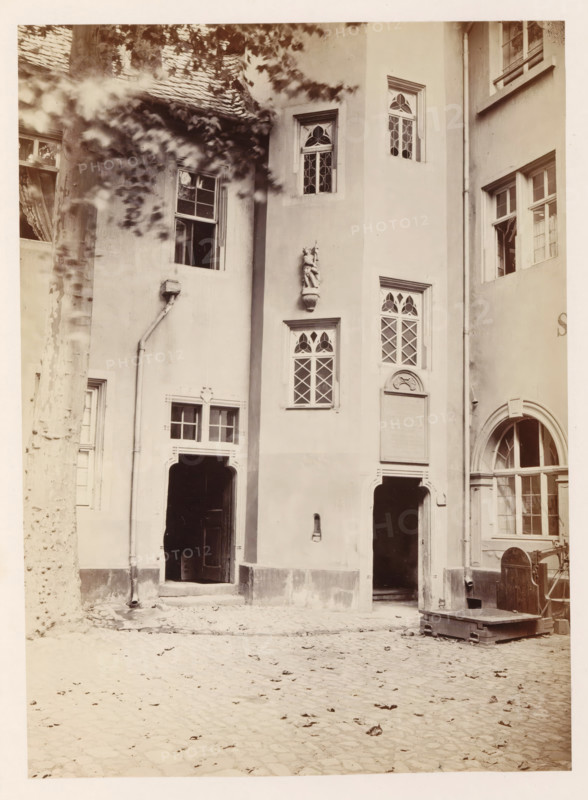 Exterior of the Zum Humbrecht house in Mainz, photograph, page, number: '18', photographer: B. Erdmann, Mainz, c. 1867 - in or before 1872, Rhineland-Palatinate, paper, albumen print, height 223 mm x width 164 mm, housing, façade (of house or building), A sepia-toned photograph of a courtyard façade showing several arched and rectangular doorways and windows set in smooth plaster walls, with decorative carved window frames and diamond-patterned leaded panes; a sculpted figure mounted on the wall above a small window; a drainpipe running down the wall beside a tall tree trunk with dappled light and blurred leaves in the foreground; cobblestone ground with scattered leaves; an open dark doorway leading into shadow, another arched entrance with panes of glass, and a wooden platform or lift with metal fittings positioned near the right side.