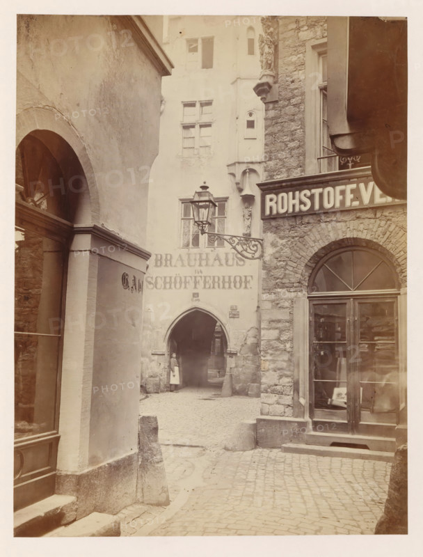 Exterior of Peter Schöffer's printing house in Mainz, photograph, page, number: '19', photographer: B. Erdmann, Mainz, c. 1867 - in or before 1872, B. Erdmann, photographer, Rhineland-Palatinate, paper, albumen print, height 235 mm x width 177 mm, printing, printing office, façade (of house or building), A narrow cobblestone alley flanked by textured stone and plaster buildings with arched doorways and windows, a decorative wrought iron street lamp projecting from a wall, a carved stone figure mounted on a corner, a glass storefront with wooden framing and visible interior shelves, a rounded arch passage leading further into the alley where a person in a light apron stands near the entrance, steps and low stone bollards at the walkway edges, and overhanging architectural elements casting soft shadows across the scene.