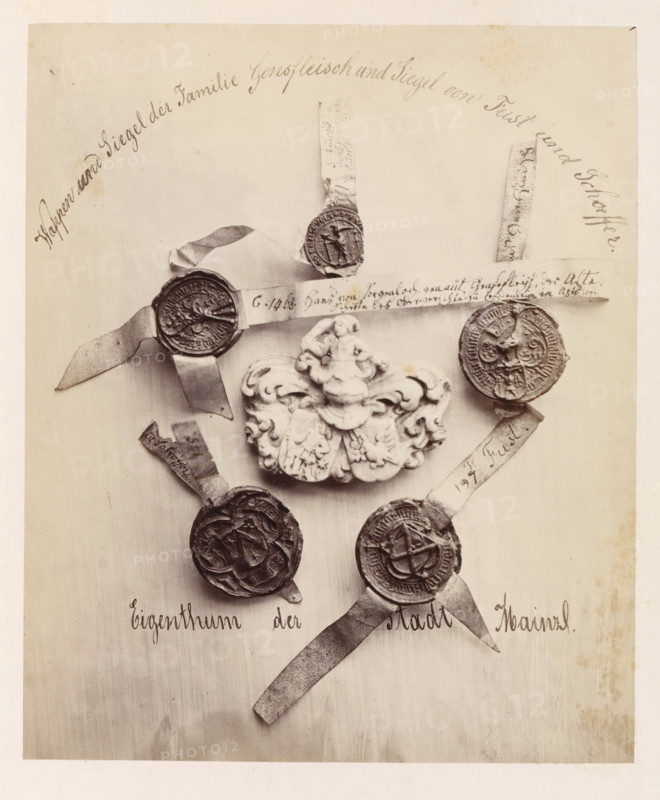 Family coat of arms and seal of the Gensfleisch family and seals of the Fust and Schöffer families, photograph, page, number: '30', photographer: B. Erdmann, Mainz, c. 1867 - in or before 1872, Rhineland-Palatinate, paper, albumen print, albumendruk, height 197 mm x width 164 mm, armorial bearing, heraldry, seal, stamp, A sepia-toned photograph shows several round wax seals attached to narrow strips of paper arranged around an ornate carved crest. The crest is sculpted with flowing scrollwork and a central raised figure. The seals vary in design and texture, some showing raised emblems or figures and others with complex patterned borders; each is affixed to a frayed paper tail. The seals and crest are displayed against a plain light background with visible faint stains and slight discoloration near the edges. There is decorative cursive writing along the top and bottom margins of the image.