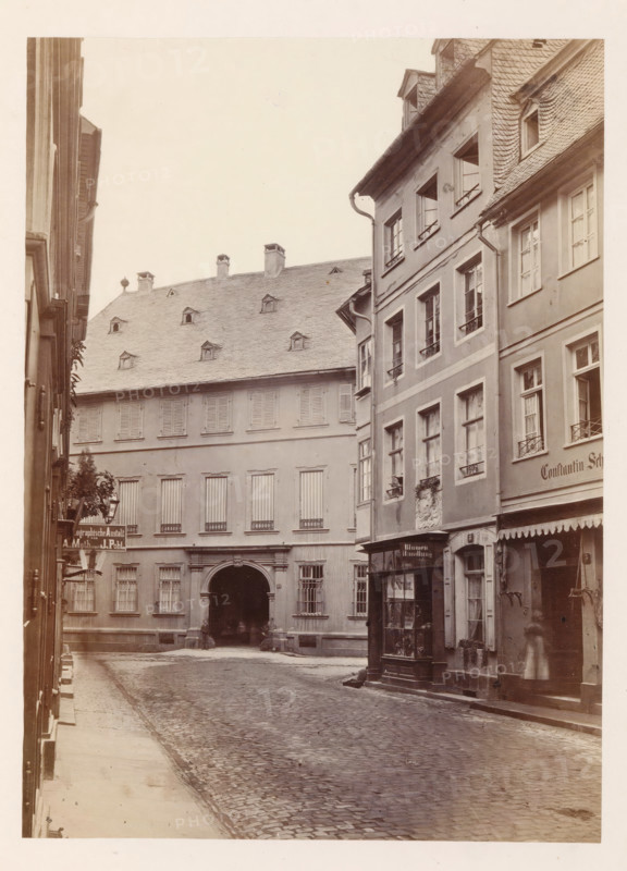Exterior of the home of the Gensfleisch family in Mainz, photograph, page, number: '4', photographer: B. Erdmann, Mainz, c. 1867 - in or before 1872, Rhineland-Palatinate, paper, albumen print, height 207 mm x width 150 mm, housing, façade (of house or building), A narrow cobblestone street flanked by multi-story buildings with shuttered and paned windows, a recessed arched passageway in the central building, steeply pitched roofs with small dormer windows and chimneys, a storefront with a glass display and an awning on the right, decorative window boxes and railings, a drainpipe running down the facade, and a faint blurred figure near the right doorway.