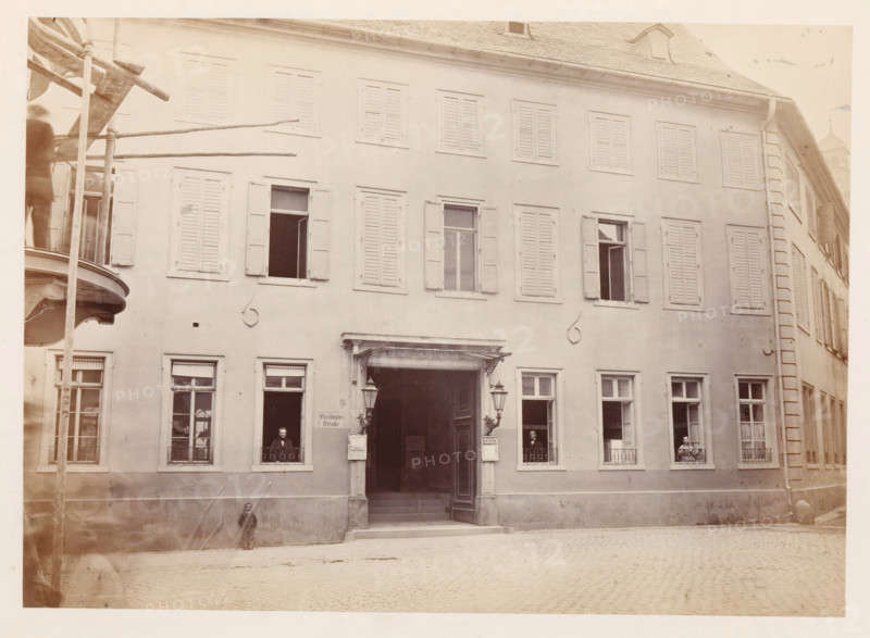 Front facade of the Gensfleisch family home in Mainz, photograph, page, number: '5', photographer: B. Erdmann, Mainz, c. 1867 - in or before 1872, Rhineland-Palatinate, paper, albumen print, height 173 mm x width 240 mm, housing, façade (of house or building), A multi-story building façade with rows of shuttered windows and a central open doorway flanked by decorative lamps, a small canopy above the entrance, and railings at the lower windows; several open windows reveal seated figures visible through the frames, a person standing near the left entrance window and another near the right; a curved balcony structure and vertical wooden poles are visible at the far left edge, and a cobblestone street runs along the front of the building.