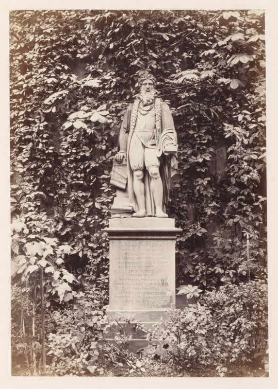 Statue of Johannes Gutenberg in Mainz, photograph, page, number: '6', photographer: B. Erdmann, Mainz, c. 1867 - in or before 1872, Rhineland-Palatinate, paper, albumen print, height 197 mm x width 140 mm, sculpture, A stone statue of a bearded man stands on a rectangular plinth surrounded by dense foliage; the figure wears draped clothing with a long braided element over the shoulders and holds a folded scroll in one hand while a book rests near the other hand, the statue's legs are exposed below knee-length trousers and the base shows weathered surfaces, all set against a backdrop of climbing leaves and plants with foliage extending around and above the monument.