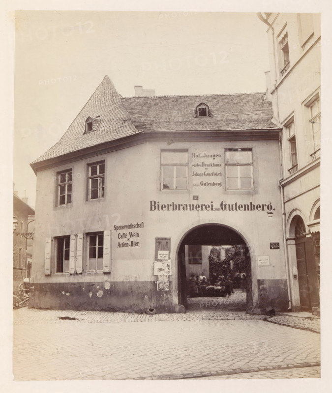 Exterior of the former printing house of Johannes Gutenberg in Mainz, photograph, page, number: '7', photographer: B. Erdmann, Mainz, c. 1867 - in or before 1872, Rhineland-Palatinate, paper, albumen print, height 205 mm x width 171 mm, printing, printing office, façade (of house or building), A sepia photograph of an old two-story building with a steep tiled roof and small dormer windows, showing weathered plaster walls, shuttered windows on the left, and an arched passageway through the center leading to a courtyard where a group of people are seated; posters and notices are pasted beside the arch, the building façade shows patches of peeling plaster and stains, cobblestone pavement fills the foreground, and adjacent multi-story buildings frame the right side of the image.