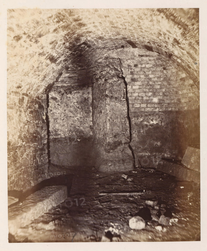 Interior of the former printing house of Johannes Gutenberg in Mainz, photograph, page, number: '9', photographer: B. Erdmann, Mainz, c. 1867 - in or before 1872, Rhineland-Palatinate, paper, albumen print, albumendruk, height 217 mm x width 181 mm, printing, printing office, A dimly lit underground space with a low arched ceiling made of rough bricks and mortar, a central vertical column of stone showing a large irregular crack, uneven stone walls on either side with visible mortar lines, a rough stone floor strewn with scattered debris and a few larger flat stones, and areas of light and shadow emphasizing the textured, aged surfaces.