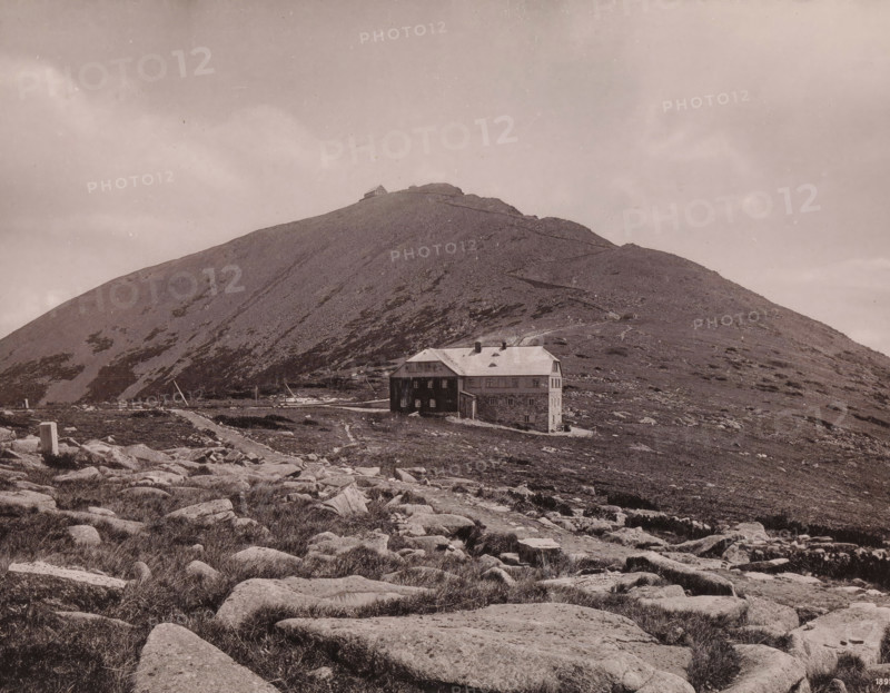 Die Schneekoppe mit der Riesenbaude, View of the Schneekoppe, page, photomechanical print, maker: E. Mertens & Cie, Giant Mountains, publisher: Max Leipelt, Warmbrunn, 1891, Silesia, Dolnoslaskie voivodship, Jelenia Góra, paper, collotype, photoprint, height 202 mm x width 268 mm, mountains, housing, façade (of house or building), Schneekoppe, A wide landscape with a rocky foreground and scattered low vegetation leading toward a gently sloping hillside, a large multi-story stone and wooden building with a pitched roof sits near the base of the hill, a narrow winding path ascends the hill toward a small structure near the summit, the hillside shows patches of bare rock and sparse low plants, the sky above is pale and mostly uniform with faint cloud formations.