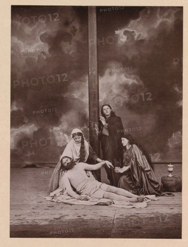 Pietà, page, photomechanical print, photographer: Carl Stockmann, Oberammergau, cliché maker: anonymous, c. 1885 - in or before 1890, Bavaria, paper, collotype, photoprint, height 224 mm x width 166 mm, 'Pietà ', 'Vesperbild', 'Marienklage' (no others present): Christ, either with or without crown of thorns, mourned by Mary, group of actors, troupe, actors on the stage, A sepia-toned staged scene showing a horizontal wooden beam or crosspiece rising from the floor toward the top, set against a cloudy backdrop. A shirtless man lies on the floor draped with a cloth, his head tilted back and eyes closed, with a beard and long hair. A woman wearing a light-colored head covering and cloak kneels behind him, supporting his head and looking downward. Another woman in darker drapery kneels at his side, holding his hand and gazing toward him. A standing figure in a dark cloak leans against the vertical beam, hands clasped and looking upward. A small covered vessel sits on a rounded stone to the...