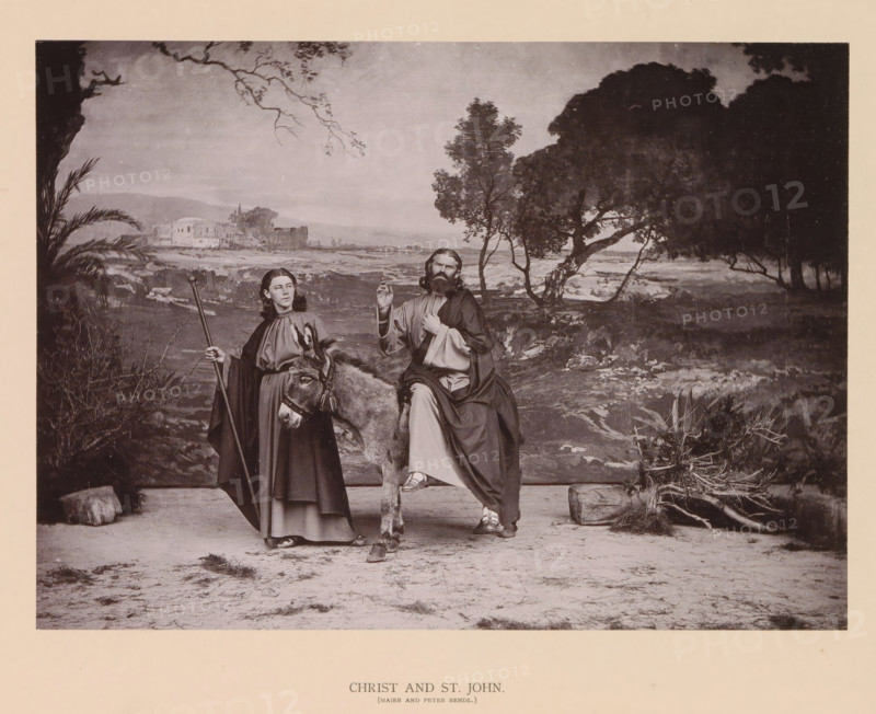 Christ and St. John, page, photomechanical print, inscription: 'Gemeinde Oberammergau Passionsspiel 1890', photographer: Carl Stockmann, Oberammergau, cliché maker: anonymous, c. 1885 - in or before 1890, Bavaria, paper, collotype, photoprint, height 167 mm x width 227 mm, Person, Jos Maier, Peter Rendl, portrait of actor, actress, Christ, life and acts of John the Evangelist, A sepia-toned scene shows two robed figures and a donkey in a landscape with distant buildings and trees; one figure stands holding a staff and the donkey’s harness while wearing a long robe and cloak, the other is seated on the donkey with a beard and long hair, draped in layered robes and raising one hand; the donkey faces forward with visible bridle details and textured fur; the ground in front is bare and sandy with scattered vegetation and rocks, while the background includes rolling terrain, a cluster of pale buildings, scattered trees with branching limbs, and a darker mass of foliage to the right, all fra...