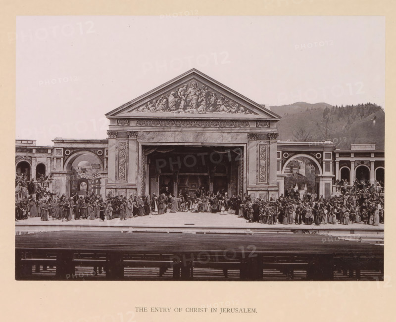 The entry of Christ in Jerusalem, page, photomechanical print, inscription: 'Gemeinde Oberammergau Passionsspiel 1890', photographer: Carl Stockmann, Oberammergau, cliché maker: anonymous, c. 1885 - in or before 1890, Bavaria, paper, collotype, photoprint, height 166 mm x width 229 mm, group of actors, troupe, actors on the stage, entry into Jerusalem: people spreading their clothes before Christ on the ass, and waving palm branches (Matthew 21:1-11, 21:14-16, Mark 11:1-11, Luke 19:28-40, John 12:12-19), A wide stage set resembling a classical temple façade with a large central pediment showing sculpted figures above a deep recessed archway, flanked by ornate columns and decorative panels, with side arches and architectural backdrops extending left and right; a dense assembly of people in period clothing fills the stage area, grouped in clusters and facing various directions, with some figures on elevated steps and others standing in front of the central opening; rows of benches or sea...