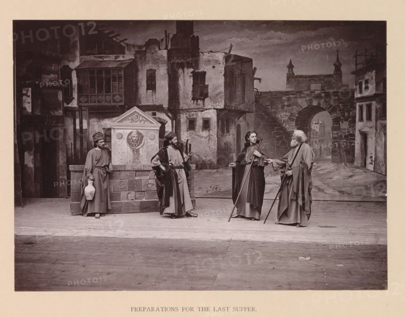 Preparations for the last supper, page, photomechanical print, inscription: 'Gemeinde Oberammergau Passionsspiel 1890', photographer: Carl Stockmann, Oberammergau, cliché maker: anonymous, c. 1885 - in or before 1890, Bavaria, paper, collotype, photoprint, height 168 mm x width 227 mm, group of actors, troupe, actors on the stage, the episode of the Last Supper, A staged scene shows four figures in long robes standing on a wooden floor in front of a painted backdrop of an old stone street with buildings and an arched gateway; two of the figures hold long staffs, one holds a jug, and another rests a hand on a stone fountain structure that has a carved face relief; the figures wear head coverings or cloaks and have varied facial hair and hairstyles, while the backdrop depicts textured walls, shuttered windows, and architectural details such as balconies and stone steps under a cloudy sky.