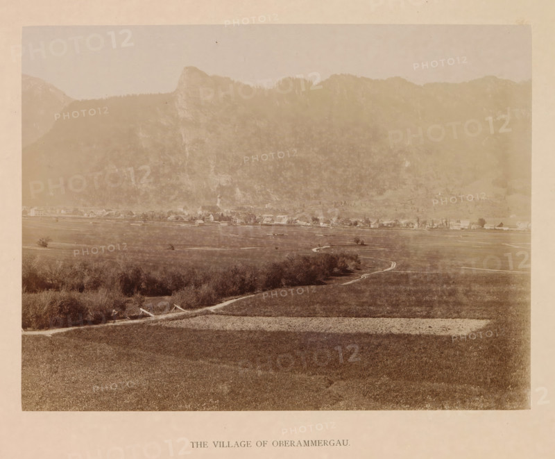 The village of Oberammergau, photograph, page, inscription: 'G. Stuffler K. Hofkunsthandlung Munich', Bavaria, c. 1885 - in or before 1890, photographer, paper, albumen print, height 168 mm x width 220 mm, city-view in general, 'veduta', Oberammergau, A wide sepia-toned landscape photograph showing a flat grassy foreground with a winding dirt path leading toward a distant village, low shrubs and small clusters of trees scattered along the field, a line of buildings with pitched roofs and a church steeple at the village edge, beyond the village a steep wooded hillside rising into craggy mountain peaks, and a pale sky above.