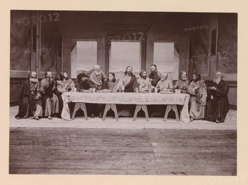 The holy supper, page, photomechanical print, inscription: 'Gemeinde Oberammergau Passionsspiel 1890', photographer: Carl Stockmann, Oberammergau, cliché maker: anonymous, c. 1885 - in or before 1890, Bavaria, paper, collotype, photoprint, height 165 mm x width 227 mm, group of actors, troupe, actors on the stage, the episode of the Last Supper, A long rectangular scene shows a group of people seated closely along one side of a long table draped with a patterned cloth, with wooden trestle legs visible beneath; the table holds plates of food, cups, and a few vessels. The people wear long robes and have varied facial hair and hairstyles, with some looking toward each other and others facing forward; one person stands behind the table leaning toward the seated figures. Behind them are tall framed wall panels with painted landscape inserts and darker surrounding walls with vertical pilasters. The floor in front of the table is wooden planks extending toward the foreground. The image is mon...
