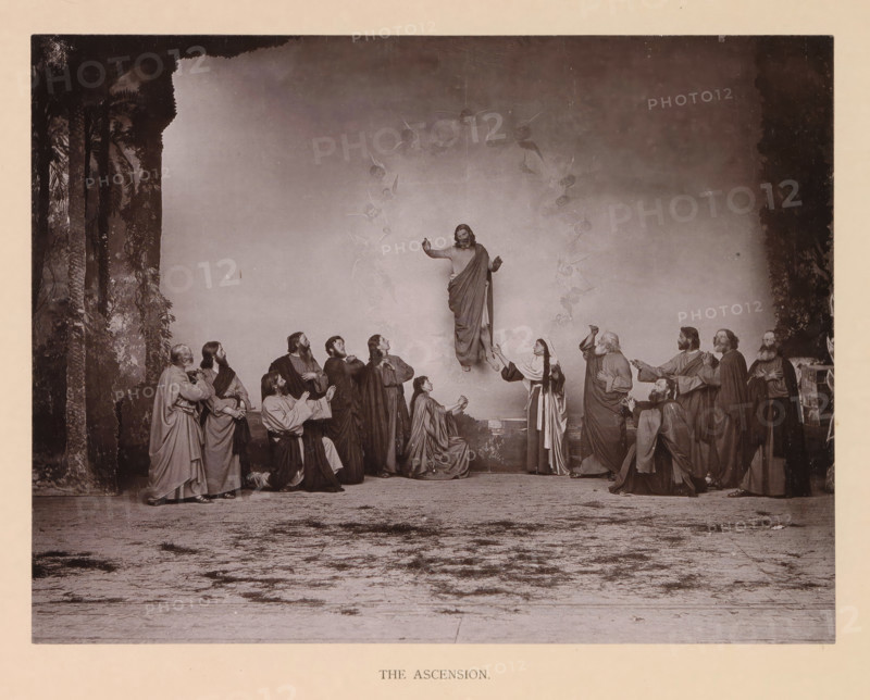 The ascension, page, photomechanical print, inscription: 'Gemeinde Oberammergau Passionsspiel 1890', photographer: Carl Stockmann, Oberammergau, cliché maker: anonymous, c. 1885 - in or before 1890, Bavaria, paper, collotype, photoprint, height 176 mm x width 230 mm, Print after page 132., group of actors, troupe, actors on the stage, Christ aloft in the sky, Ascension, A sepia-toned scene shows a robed figure elevated above a group, with arms raised and draped cloth flowing around the body, surrounded by faint winged figures forming a vertical arc; below, a circle of men and women in long robes stand, kneel, or reach upward with varied gestures and facial expressions, some looking up and some holding hands, arranged across a stage-like ground with a textured foreground; tall dark foliage frames the left and right edges of the scene, creating a backdrop against a plain wall behind the central figures.