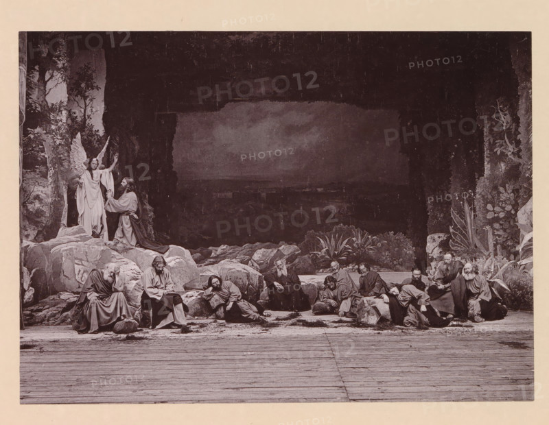 Christ's agony in the garden of Gethsemani, page, photomechanical print, inscription: 'Gemeinde Oberammergau Passionsspiel 1890', photographer: Carl Stockmann, Oberammergau, cliché maker: anonymous, c. 1885 - in or before 1890, Carl Stockmann, photographer, cliché maker, Bavaria, paper, collotype, photoprint, height 167 mm x width 227 mm, group of actors, troupe, actors on the stage, agony of Christ: to comfort him one or more angels appear to Christ with chalice and, or cross, A wide scene showing a group of robed men gathered among large rocks and plants in front of a dark cave opening, with some men seated, some reclining, and some leaning on stones; an angelic figure with wings stands on a rock near the cave entrance raising one hand while a bearded man kneels before the angel; surrounding the central figures are rocky formations, foliage, and a wooden floor in the foreground.