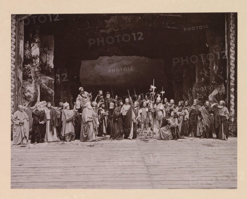 The betrayal, page, photomechanical print, inscription: 'Gemeinde Oberammergau Passionsspiel 1890', photographer: Carl Stockmann, Oberammergau, cliché maker: anonymous, c. 1885 - in or before 1890, Bavaria, paper, collotype, photoprint, height 162 mm x width 208 mm, group of actors, troupe, actors on the stage, the kiss of Judas: accompanied by soldiers with torches and lanterns, he kisses Christ, A wide stage scene framed by patterned vertical borders shows a group of people dressed in long robes and period garments gathered across the middle ground, some standing and some seated, with a few holding long pole weapons and banners; the background is dark with painted foliage and rock formations at the sides, and the foreground is a wooden floor with visible planks extending toward the viewer.