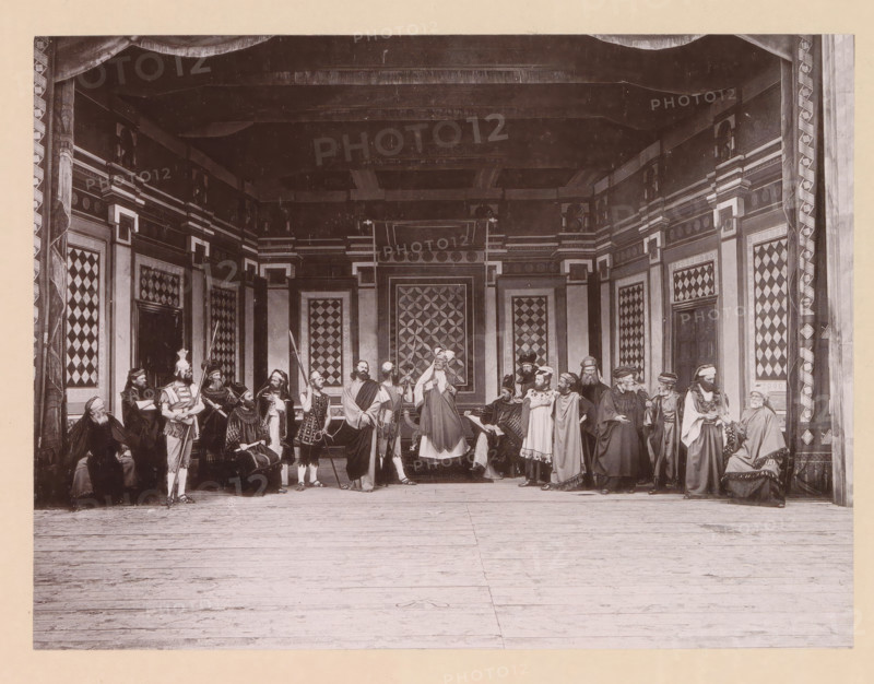 Christ before the highpriest Caiaphas, page, photomechanical print, inscription: 'Gemeinde Oberammergau Passionsspiel 1890', photographer: Carl Stockmann, Oberammergau, cliché maker: anonymous, c. 1885 - in or before 1890, Bavaria, paper, collotype, photoprint, height 162 mm x width 216 mm, group of actors, troupe, actors on the stage, Christ before the Sanhedrin with Caiaphas as high priest, and possibly Annas, maybe a soldier about to strike Christ because he keeps silent (Matthew 26:57-66, Mark 14:53-64, Luke 22:54, 22:66-71), A wide stage scene shows a group of people in historical costumes standing and sitting across an ornate set with patterned wall panels and decorative columns, a draped proscenium at the top, and a wooden floor in the foreground; the figures wear robes, tunics, helmets, cloaks, and head coverings, and some hold staffs, spears, or other props while others are seated or gesturing, with detailed geometric designs visible on the backdrop and side panels.
