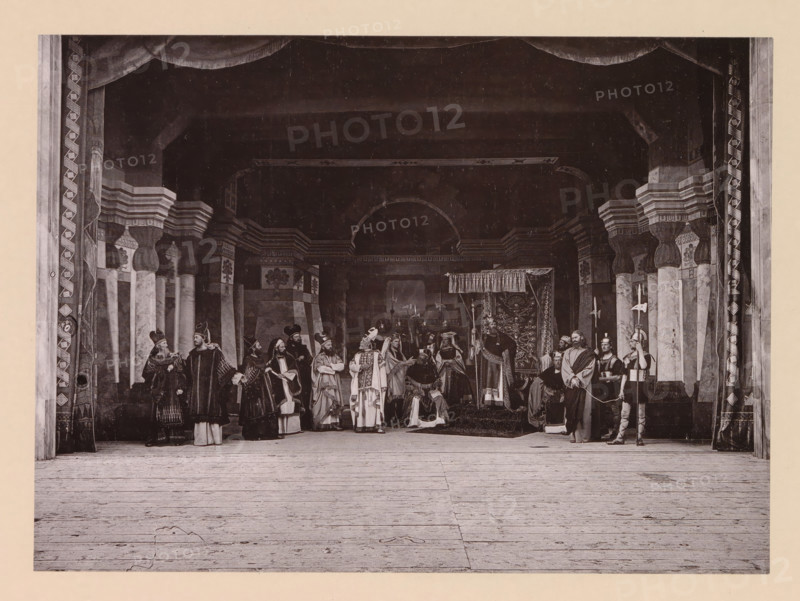 Christ before king Herod, page, photomechanical print, inscription: 'Gemeinde Oberammergau Passionsspiel 1890', photographer: Carl Stockmann, Oberammergau, cliché maker: anonymous, c. 1885 - in or before 1890, Bavaria, paper, collotype, photoprint, height 165 mm x width 226 mm, group of actors, troupe, actors on the stage, sometimes a gorgeous robe is brought (Luke 23:8-11), A wide stage set with tall decorative columns and an arched backdrop, draped curtains framing the scene, and a wooden floor in the foreground; a group of people in historical costumes stand and sit across the stage, wearing robes, capes, headdresses, and armor, some holding staffs, spears, banners, or other props, with a seated figure beneath a canopy toward the center and patterned tapestries or decorative panels behind them.