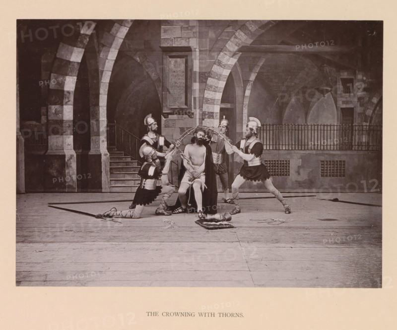 The crowning with thorns, page, photomechanical print, inscription: 'Gemeinde Oberammergau Passionsspiel 1890', photographer: Carl Stockmann, Oberammergau, cliché maker: anonymous, c. 1885 - in or before 1890, Bavaria, paper, collotype, photoprint, height 167 mm x width 227 mm, group of actors, troupe, actors on the stage, the crowning with thorns: soldiers with sticks place a thorny crown on Christ's head and give him a reed (Matthew 27:27-31, Mark 15:16-20, John 19:2-3), A staged scene set against a stone arcade with striped arches and a stairway shows a seated man with bare torso and draped cloak surrounded by four armored figures wearing helmets and tunics; two of the armored figures are kneeling while the others stand, and they hold a circular object above the seated man's head and ropes or cords around his arms, with additional props including a helmet on the floor and a cushion nearby, all on a wooden floor in front of a railing and masonry backdrop.