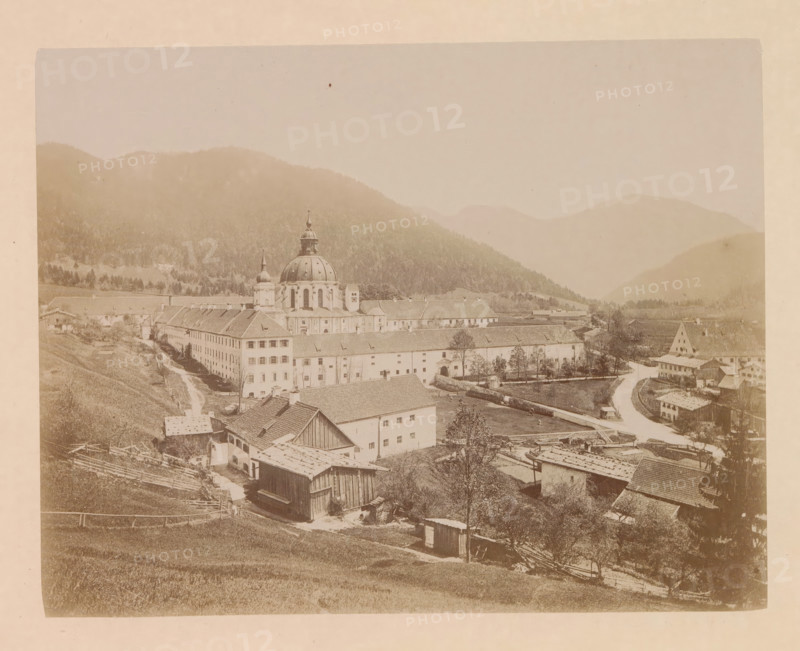 The village of Ettal, photograph, page, inscription: 'G. Stuffler K. Hofkunsthandlung Munich', Bavaria, c. 1885 - in or before 1890, photographer, paper, albumen print, height 167 mm x width 212 mm, city-view in general, 'veduta', Ettal, A sepia-toned panoramic photograph shows a valley town with a large domed church and adjacent buildings occupying the central area, surrounded by rolling hills and distant mountains shrouded in haze. The foreground contains sloping grassy fields with wooden fences, small sheds, and clusterings of rooftops and barns. A winding road curves through the scene past rows of multi-story rectangular buildings with pitched roofs and numerous windows, small trees dot the grounds, and a river or canal runs alongside part of the built area. The church features a prominent rounded dome topped by a smaller cupola and flanked by a slender tower, and terraced buildings extend from it in linear blocks. The overall image has a faded, aged appearance with soft contrast a...