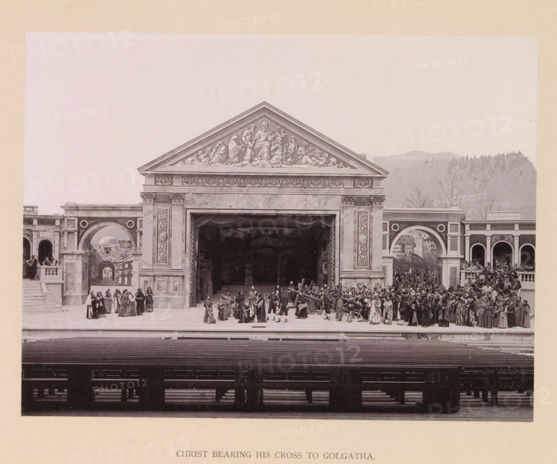 Christ bearing his cross to Golgatha, page, photomechanical print, inscription: 'Gemeinde Oberammergau Passionsspiel 1890', photographer: Carl Stockmann, Oberammergau, cliché maker: anonymous, c. 1885 - in or before 1890, Bavaria, paper, collotype, photoprint, height 167 mm x width 223 mm, group of actors, troupe, actors on the stage, from Pilate's palace to Golgotha: the procession to Calvary (Matthew 27:31-33, Mark 15:20-22, Luke 23:26-33, John 19:17), A wide stage set resembling a classical stone building with a central pediment decorated with carved figures and ornate frieze sits against a pale background with distant hills; a large open archway leads into a dark interior, flanked by tall columns with decorative panels and arches on either side. On the stage, many people in period costumes are arranged in clustered groups across the foreground, some standing on steps and balconies, others gathered near the central opening; several figures carry a large wooden beam across their shou...