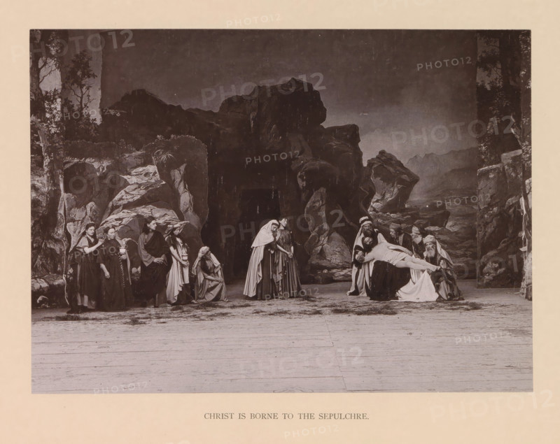 Christ is borne to the sepulchre, page, photomechanical print, inscription: 'Gemeinde Oberammergau Passionsspiel 1890', photographer: Carl Stockmann, Oberammergau, cliché maker: anonymous, c. 1885 - in or before 1890, Bavaria, paper, collotype, photoprint, height 168 mm x width 230 mm, Print after page 184., group of actors, troupe, actors on the stage, bearing of Christ's body to the grave, A sepia-toned stage scene shows a rocky backdrop with cliffs and sparse foliage framing a group of people dressed in historical robes and head coverings; on the left a line of figures stand and sit near rock formations, some with cloaks and shawls, one holding an object, several with downcast expressions, while toward the center two women in long garments stand close together and another kneels nearby; on the right a group of men with turbans and beards cradle a limp, draped body across their arms as others gather around, the foreground revealing a wide wooden stage floor and the entire composition...