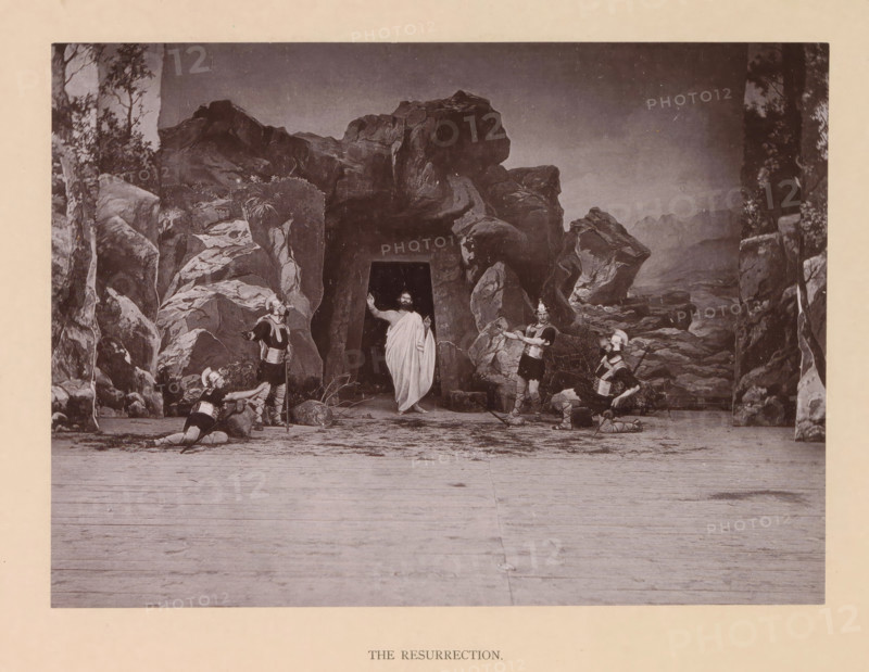 The resurrection, page, photomechanical print, inscription: 'Gemeinde Oberammergau Passionsspiel 1890', photographer: Carl Stockmann, Oberammergau, cliché maker: anonymous, c. 1885 - in or before 1890, Bavaria, paper, collotype, photoprint, height 168 mm x width 228 mm, group of actors, troupe, actors on the stage, Resurrection of Christ, A staged scene set against a painted rocky backdrop with a central dark cave entrance, a robed figure standing in the cave entrance with one arm raised, four armored figures around the cave wearing helmets and tunics with greaves, two of the armored figures kneeling and two standing, scattered rocks and low vegetation at the cave mouth, a wide wooden floor in the foreground, and tall painted rock formations and trees framing the scene on both sides.