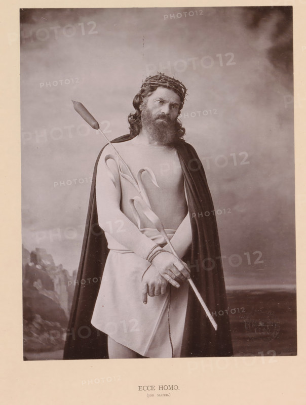 Ecce Homo (Jos Maier), page, photomechanical print, inscription: 'Gemeinde Oberammergau Passionsspiel 1890', photographer: Carl Stockmann, Oberammergau, cliché maker: anonymous, c. 1885 - in or before 1890, Bavaria, paper, collotype, photoprint, height 200 mm x width 157 mm, Person, Jos Maier, portrait of actor, actress, Christ alone (also called 'Ecce Homo'), A sepia-toned portrait of a bearded man wearing a crown of thorns and a dark cloak draped over light clothing, holding a long staff with a bulbous tip and a stem with long leaves, his hands bound with rope at the wrists and positioned in front of his torso, his head slightly tilted and eyes looking forward against a plain, softly textured backdrop with a faint suggestion of rocky shapes at the lower left.