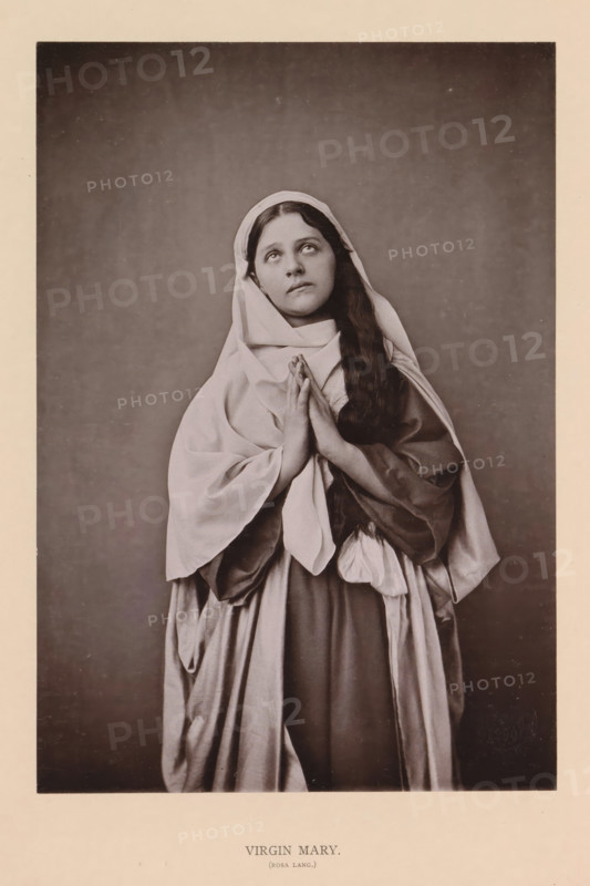 Virgin Mary (Rosa Lang), page, photomechanical print, inscription: 'Gemeinde Oberammergau Passionsspiel 1890', photographer: Carl Stockmann, Oberammergau, cliché maker: anonymous, c. 1885 - in or before 1890, Bavaria, paper, collotype, photoprint, height 215 mm x width 150 mm, Person, Rosa Lang, portrait of actor, actress, the Virgin Mary, A sepia-toned portrait of a young woman standing against a plain dark backdrop, wearing layered draped garments and a head covering, with long hair falling over one shoulder, hands held together in front of her chest, eyes looking upward, and a calm facial expression.