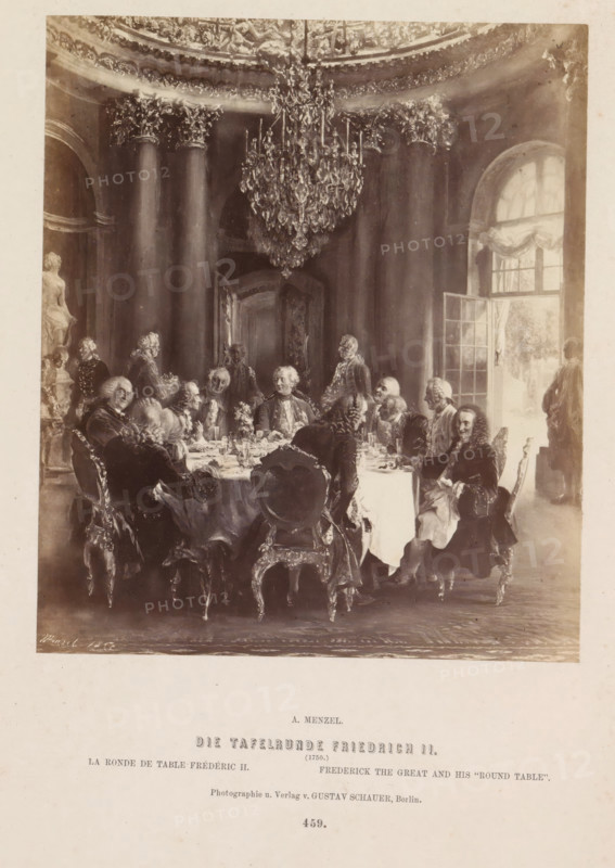 The Round Table of Frederick II, Photographic reproduction of a painting of Frederick the Great at the table in Sanssouci Palace by Adolf Menzel, photograph, reproduction, page, number: '459.', photographer: Gustav Schauer, Berlin, after painting by Adolph Menzel, c. 1858 - in or before 1868, paper, albumen print, height 165 mm x width 140 mm, Person, Frederick II (King of Prussia), evening meal, dinner, supper, table-ware, Potsdam, Sanssouci, A sepia-toned scene of a formal dining room with a large round table covered by a cloth and set with dishes and glassware, surrounded by men in period clothing with powdered wigs seated on ornate chairs; a large crystal chandelier hangs from a decorated ceiling above, fluted columns rise behind the group, a tall arched window with drapery admits light on one side, a doorway stands open in the background, a marble statue is visible near one edge, and a standing figure is near the window.