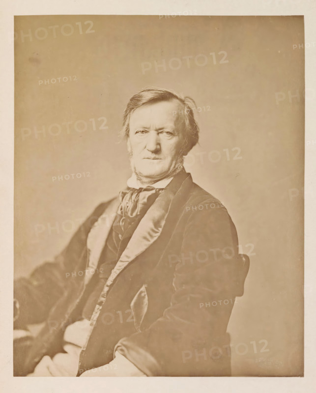 Portrait of Richard Wagner, photograph, page, photographer: Franz Hanfstaengl, Munich, c. 1873 - 1883, Bavaria, paper, albumen print, height 220 mm x width 177 mm, Photo opposite title page., Person, Richard Wagner, composer, A sepia-toned portrait of an older man seated and facing forward, showing his head and upper torso; he has light-colored hair combed to the side and sideburns, a neutral facial expression, and is wearing a dark jacket with a shiny lapel over a vest and a tied scarf at the neck; a plain, softly textured backdrop fills the space behind him and the image has a wide margin around the portrait.