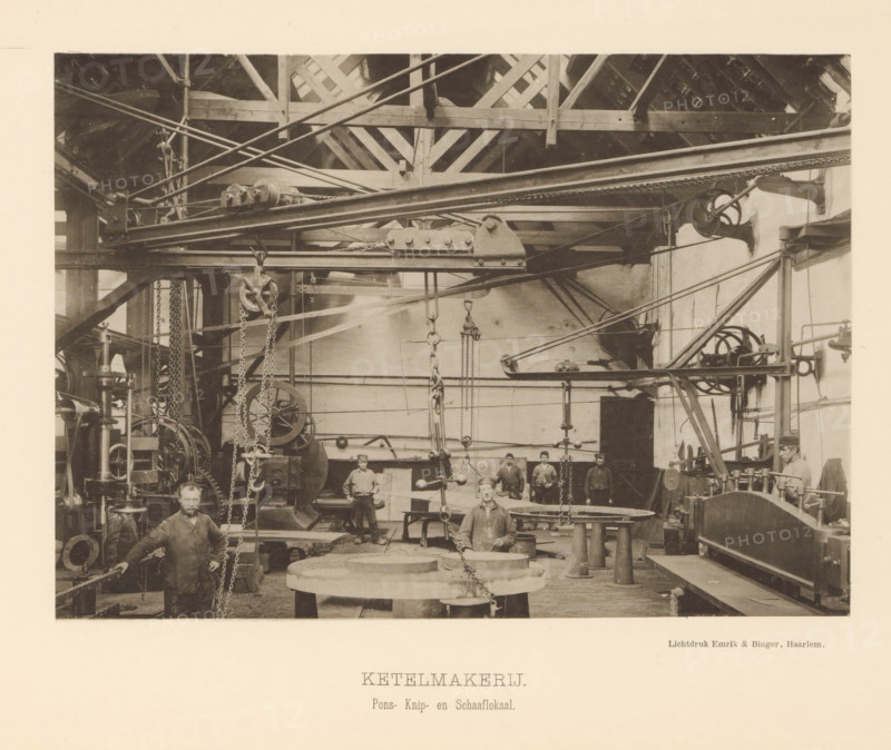 Interior with employees of the boiler shop of the machine factory of Gebroeders Stork & Co, Boiler Shop. Punching, Cutting and Planing Room, page, photomechanical print, number: 'No. 11.', Hengelo (Overijssel), maker: Emrik & Binger, Haarlem, c. 1884 - in or before 1894, photographer, paper, collotype, light print, height 155 mm x width 217 mm, factory-building, factory-interior, machine shop, A sepia-toned photograph shows an industrial interior with exposed wooden roof beams and a network of overhead metal rails, pulleys, chains, and beams. Large machinery, gears, and hanging hooks occupy the space, with round metal plates and cylindrical supports on the floor. Several men in work clothing stand or sit among the equipment, one man positioned at the front near a large circular workpiece and others grouped further back. The walls are plain and show shafts and mounted wheels, and various rods and belts run across the scene connecting machines. The image has a pale border surrounding the...