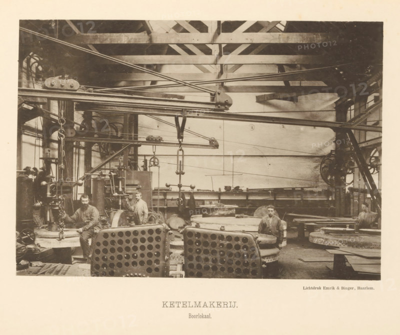 Boiler shop. Drilling room, Interior with employees of the boiler shop of the machine factory of Gebroeders Stork & Co, page, photomechanical print, number: 'No. 12.', Hengelo (Overijssel), maker: Emrik & Binger, Haarlem, c. 1884 - in or before 1894, photographer, paper, collotype, photogravure, height 152 mm x width 217 mm, factory-building, factory-interior, machine shop, A sepia-toned industrial interior shows large metal machinery, beams, pulleys, and overhead rods stretching across the space, with circular and perforated metal components in the foreground and heavy chains and hooks hanging from suspended fixtures; three men in work clothes are positioned among the equipment, two standing and one seated, each near different machines and parts, with visible gear wheels, cylinders, and workbenches around them; the ceiling structure consists of wooden trusses and beams, and windows and brick walls are visible at the sides and back, while various round metal plates and stacked componen...