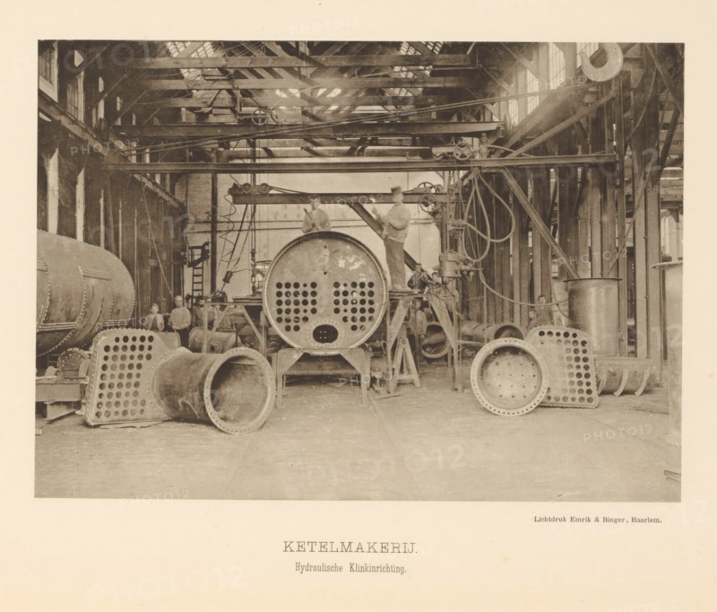 Boilerworks. Hydraulic riveting device, Interior with employees of the boiler shop of the machine factory of Gebroeders Stork & Co, page, photomechanical print, number: 'No. 13.', Hengelo (Overijssel), maker: Emrik & Binger, Haarlem, c. 1884 - in or before 1894, photographer, paper, collotype, photoprint, height 153 mm x width 216 mm, factory-building, factory-interior, machine shop, A large industrial interior with high wooden roof trusses and windows, filled with metal machinery and cylindrical boiler parts arranged across the floor, including large circular metal plates with arrays of holes and open pipe sections; scaffolding beams, pulleys, chains, and belts span the space, and several men in work clothes stand and sit on and around the machinery; vertical wooden columns line the sides and a variety of cables and mechanical linkages hang and run between beams and equipment.