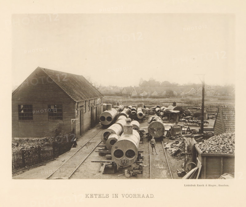 Boilers in stock, View of steam boilers at the machine factory of Stork & Co. Brothers, page, photomechanical print, number: 'No. 14.', Hengelo (Overijssel), maker: Emrik & Binger, Haarlem, c. 1884 - in or before 1894, photographer, paper, collotype, photogravure, height 156 mm x width 211 mm, factory-building, steampower (alleg.) (technical aspects, crafts, industries, agriculture), A sepia-toned scene shows an industrial yard with long cylindrical metal boilers and pipes arranged on parallel tracks, many displaying riveted circular ends, surrounded by scattered metal parts and stacks of materials; a wooden building with a pitched roof and multi-pane windows stands to the left, a narrow fenced path runs alongside it, and a man in work clothes and a hat walks between the tracks near the foreground; in the middle distance there are low houses and trees forming a horizon, and a tall pole rises near the right side of the yard.