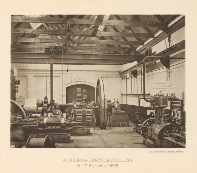 Anniversary Exhibition. 9-17 September 1893, Anniversary exhibition with steam boilers for the 25th anniversary of the machine factory of Gebroeders Stork & Co, page, photomechanical print, number: 'No. 15.', Hengelo (Overijssel), maker: Emrik & Binger, Haarlem, c. 1893-09-09 - in or before 1893-09-17, photographer, paper, collotype, photogravure, height 162 mm x width 211 mm, factory-interior, machine shop, exhibition, show, A sepia-toned interior view of a workshop showing wooden roof trusses and beams across the ceiling, a large horizontal metal girder with pulleys and chains suspended from it, and a variety of industrial machinery and mechanical equipment arranged on brick and concrete supports across the floor; a large flywheel and wheel with spokes are visible, along with pipes, belts, motors, and control components, and a pair of tall arched doors with slatted panels sit against the rear wall, flanked by brickwork and plastered walls with windows on one side.