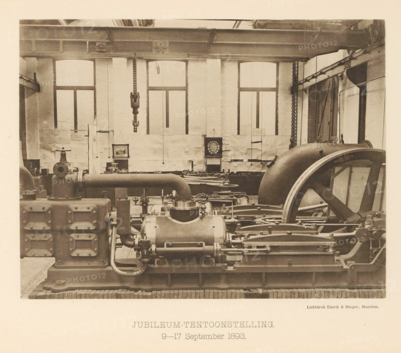 Anniversary Exhibition. 9-17 September 1893, Anniversary exhibition with a steam boiler for the 25th anniversary of the machine factory of Gebroeders Stork & Co, page, photomechanical print, number: 'No. 16.', Hengelo (Overijssel), maker: Emrik & Binger, Haarlem, c. 1893-09-09 - in or before 1893-09-17, photographer, paper, collotype, photogravure, height 165 mm x width 216 mm, factory-interior, machine shop, exhibition, show, A sepia-toned industrial interior showing large mechanical equipment and heavy machinery arranged across the floor, including a prominent flywheel with spokes on the right, large cylindrical housings and pipes in the foreground, rectangular casings with bolted access panels on the left, and various rods, linkages, and valves connecting components; the background wall features tall arched windows with panes and a brick surface, chains and hooks hanging from an overhead girder that spans the room, and a framed decorative circular object mounted on the wall among sh...