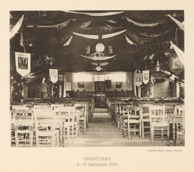 Marquee for the 25th anniversary of the machine factory of Gebroeders Stork & Co, Marquee. 9-17 September 1893, page, photomechanical print, number: 'No. 17.', Hengelo (Overijssel), maker: Emrik & Binger, Haarlem, c. 1893-09-09 - in or before 1893-09-17, photographer, paper, collotype, photogravure, height 166 mm x width 213 mm, festivities, A large indoor space arranged with rows of wooden chairs facing a raised area at the far end, the ceiling and upper walls draped with long fabric swags and foliage garlands, round hanging lanterns and decorative wreaths suspended from above, framed banners and pictorial panels hanging along the sides, tall columns or posts wrapped with greenery and floral arrangements, a central aisle leading toward a backdrop with panels and framed pictures, and wooden floorboards visible beneath the chairs.