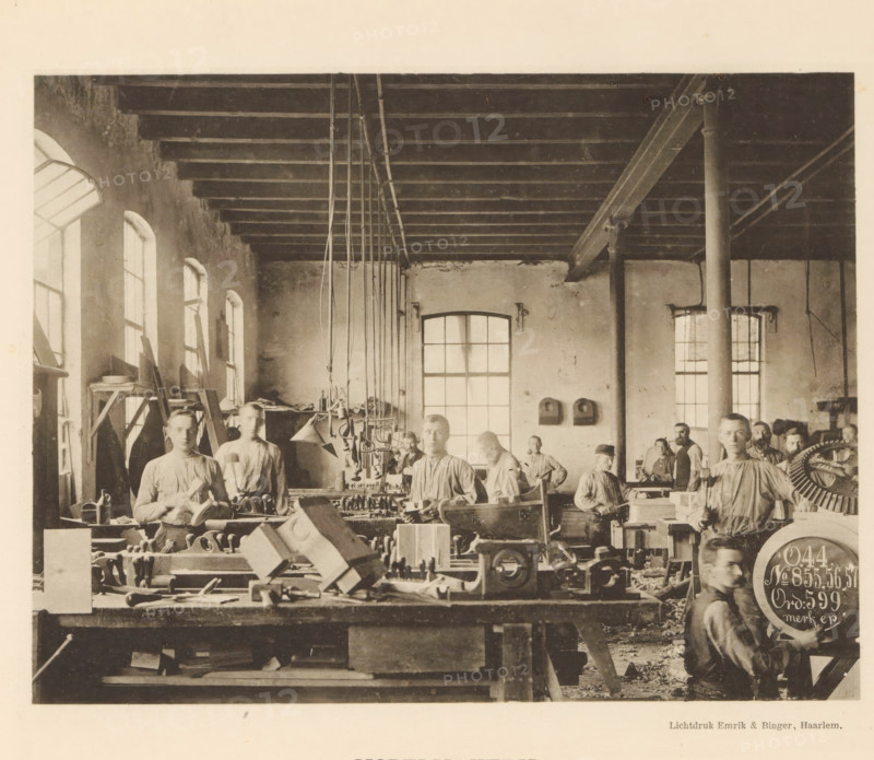 Employees making models in the machine factory of Gebroeders Stork & Co, Model making, page, photomechanical print, number: 'No. 3.', Hengelo (Overijssel), maker: Emrik & Binger, Haarlem, c. 1884 - in or before 1894, photographer, paper, collotype, photogravure, height 154 mm x width 202 mm, factory-building, workshop, A sepia-toned workshop interior showing a long room with high windows along one wall and wooden ceiling beams overhead, rows of workbenches cluttered with tools and wooden parts filling the foreground, several men in work shirts standing and sitting among the benches and machinery, hanging pipes and pulleys suspended from the ceiling, tall columns supporting a beam, and scattered shavings and materials on the floor.