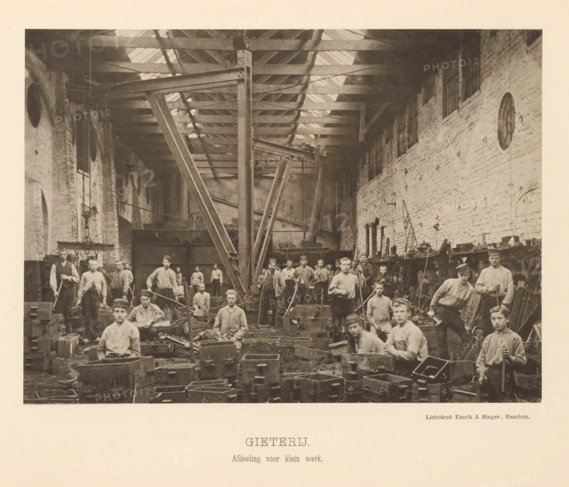 Interior with employees of the foundry of the machine factory of Gebroeders Stork & Co, Foundry. Small work department, page, photomechanical print, number: 'No. 5.', Hengelo (Overijssel), maker: Emrik & Binger, Haarlem, c. 1884 - in or before 1894, photographer, paper, collotype, photogravure, height 154 mm x width 213 mm, factory-building, workshop, A sepia-toned photograph showing the interior of a large industrial workshop with a high roof supported by visible beams and a central vertical metal column with angled braces, a long brick wall with windows and circular openings along one side, workbenches and shelves lined with tools and objects, many men and boys arranged throughout the space wearing work clothes and caps, several standing and several seated among wooden and metal molds and boxes on the floor, workshop equipment such as cranes, hoists, and large metal structures in the background, and scattered tools and material pieces across the work area.