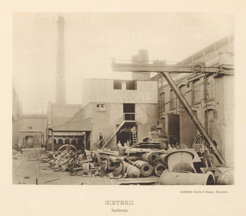 Foundry. Smelting furnaces, View of the smelting furnaces of the foundry of the machine factory of Gebroeders Stork & Co, page, photomechanical print, number: 'No. 6.', Hengelo (Overijssel), maker: Emrik & Binger, Haarlem, c. 1884 - in or before 1894, photographer, paper, collotype, photogravure, height 155 mm x width 200 mm, factory-building, fire, oven, furnace, interior of industrial structure, workshop, A sepia-toned industrial yard with piles of large metal cylinders, wheels, and assorted castings scattered across the ground in front of brick buildings; an elevated metal beam structure with a pulley wheel and supporting diagonal brace spans between buildings; a multi-story brick and corrugated structure with external stairs, ladders, and a doorway shows small groups of people standing near the entrance and on the stairs; tall chimneys and rooftop elements rise behind the buildings, and tracks and pathways run through the yard among equipment and debris.