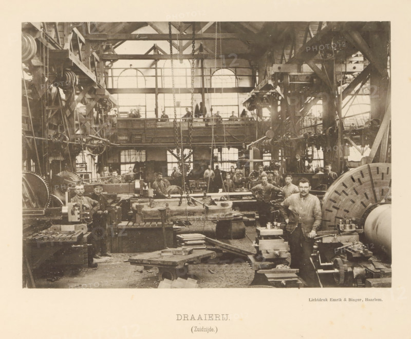 Turning shop. (South side), Interior with employees of the machine factory of Gebroeders Stork & Co, page, photomechanical print, number: 'No. 8.', Hengelo (Overijssel), maker: Emrik & Binger, Haarlem, c. 1884 - in or before 1894, photographer, paper, collotype, photogravure, height 154 mm x width 214 mm, factory-building, factory-interior, machine shop, A large industrial interior with high windows and exposed beams fills the scene, showing numerous metal machines, gears, chains, pulleys, and workbenches arranged across the floor and along raised platforms; a group of men in work clothes stand and sit among the equipment, some near a large circular gear-like wheel on the right and others by heavy blocks and machine parts in the center and left; hanging chains descend from the ceiling and scaffold-like wooden and metal structures frame the space, while scattered tools, stacked plates, and mechanical components lie on tables and the floor, and a balcony with railings runs across the bac...