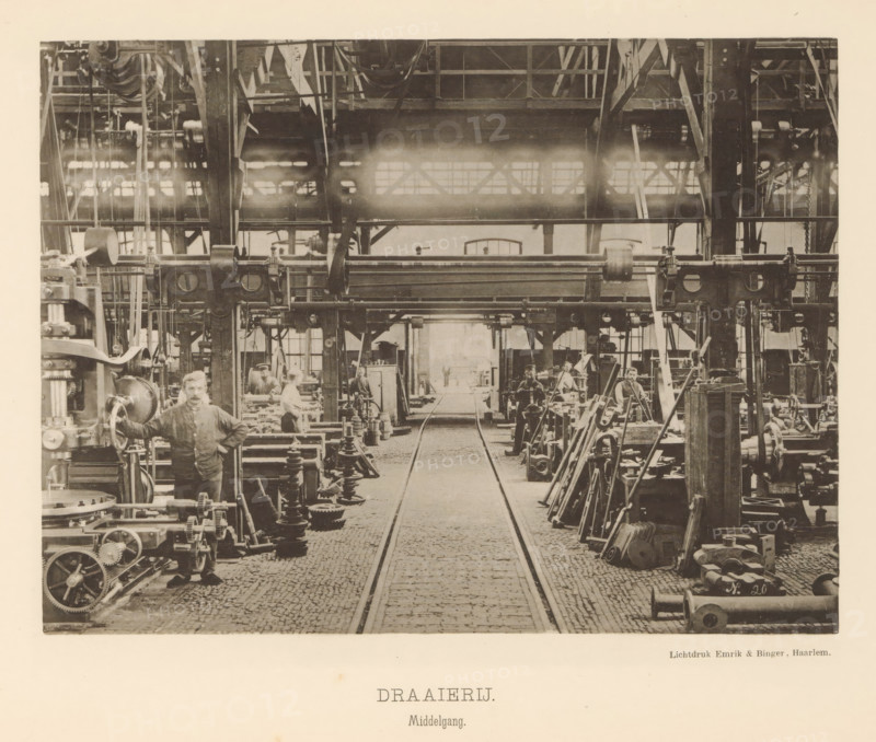 Interior with employees of the machine factory of Gebroeders Stork & Co., Turning Shop. Central aisle, page, photomechanical print, number: 'No. 9.', Hengelo (Overijssel), maker: Emrik & Binger, Haarlem, c. 1884 - in or before 1894, photographer, paper, collotype, photogravure, height 157 mm x width 210 mm, factory-building, factory-interior, machine shop, A wide interior view of an industrial workshop with a cobblestone central aisle flanked by metal tracks leading toward a distant open doorway, heavy overhead steel beams and a gallery with pulleys and belts near the ceiling, rows of large machines and gear assemblies lining both sides of the aisle, stacks of metal parts and tools arranged beside the machines, several men in work clothes standing and sitting at the machinery, a man in the foreground with one hand resting on a large machine wheel, ladders and structural columns interspersed among the equipment, and light coming through high windows near the back of the space.